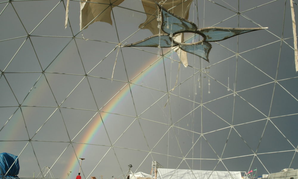 The rosewater chandelier hangs in the middle of a 50 foot geodesic dome during burning man. A daytime rainbow after a storm passes through the handblown glass