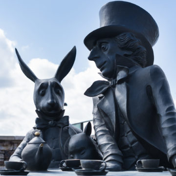 Bronze sculpture of the mad hatter and white rabbit sitting at a table having tea. Blue sky background.