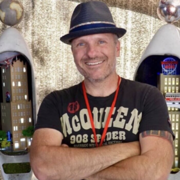 a photo of scott hildebrandt a.k.a. "Mister Christmas" standing in front of his artwork with a bowler hat with arms crossed.